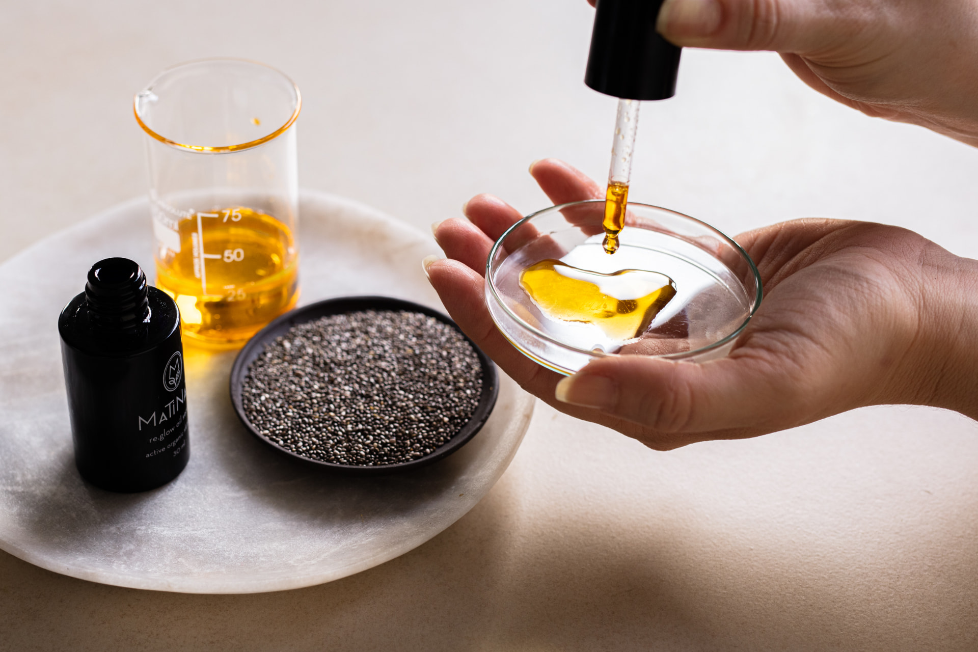 A pipette releasing golden botanical oil into a glass dish beside a Matinata bottle, a measuring beaker and chia seeds — small-batch formulation in the lab