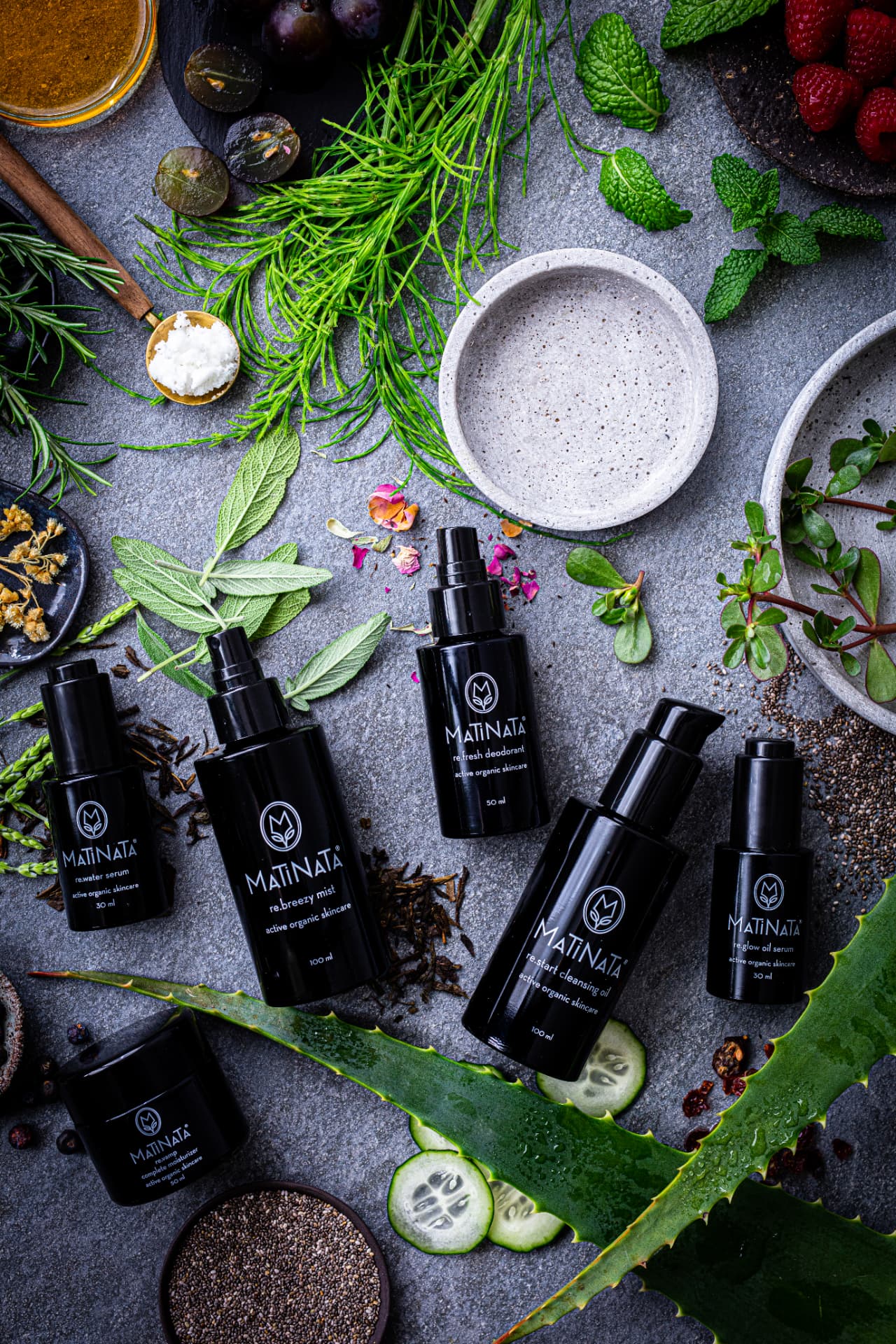 Matinata violet glass bottles surrounded by fresh rosemary, sage, mint, raspberries, cucumber and aloe on stone